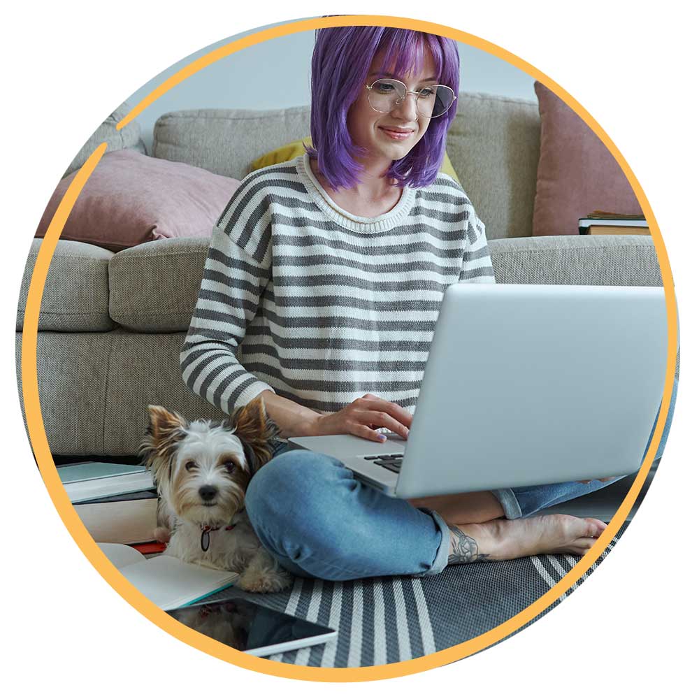 Woman with purple hair working on laptop with dog next to her